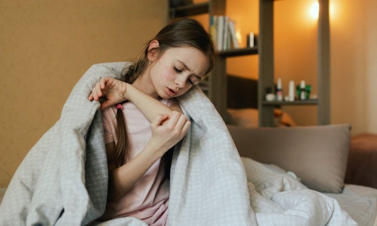 Junges Mädchen sitzt im Bett, in eine Decke gehüllt, und kratzt sich am Unterarm; im Hintergrund sind Regal und warmes Zimmerlicht zu sehen